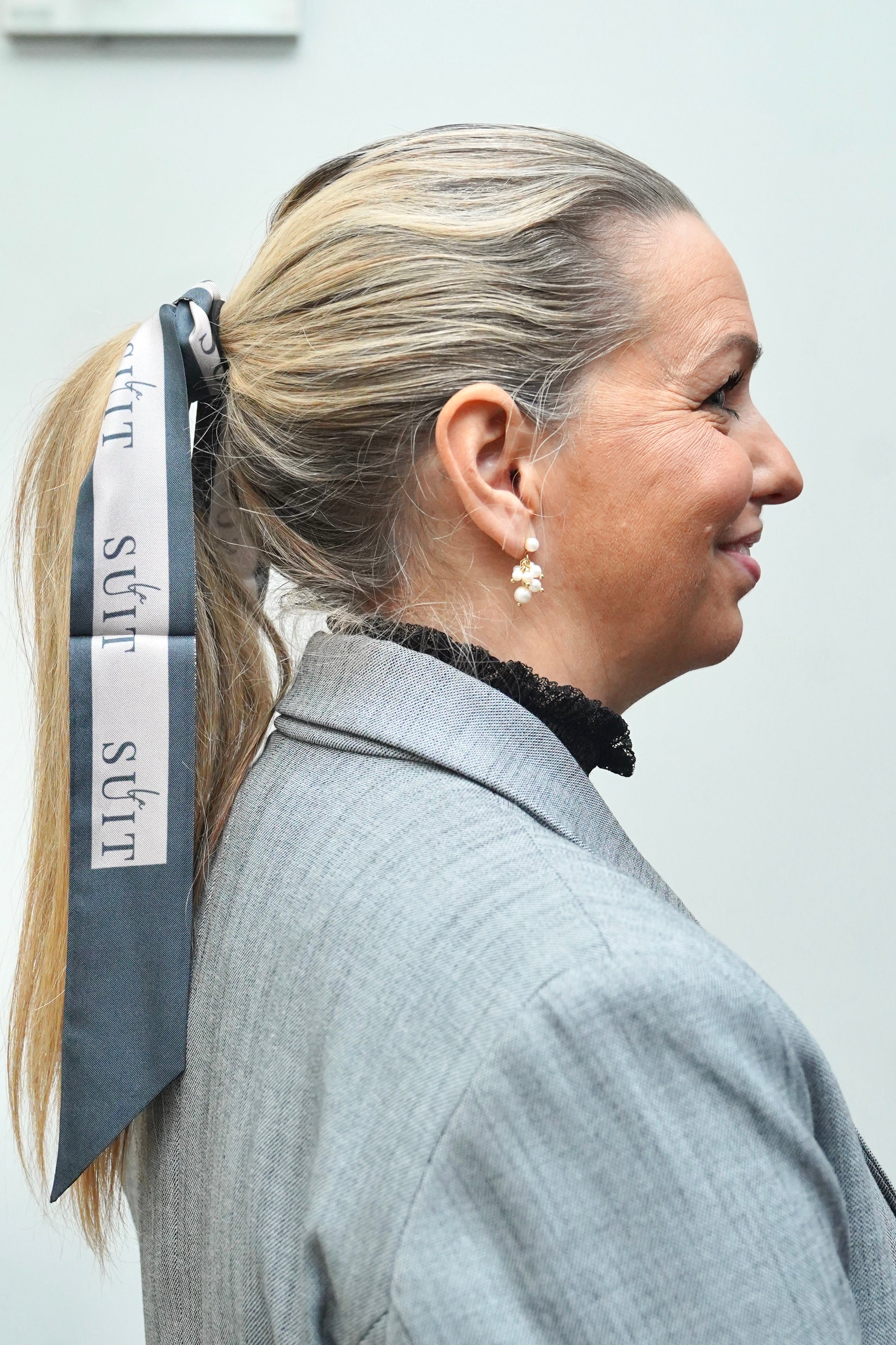 Kvinde iført LA SUIT produkter, herunder beige silke twilly i håret og grå habitjakke set fra siden.