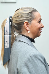 Kvinde iført LA SUIT produkter, herunder beige silke twilly i håret og grå habitjakke set fra siden.